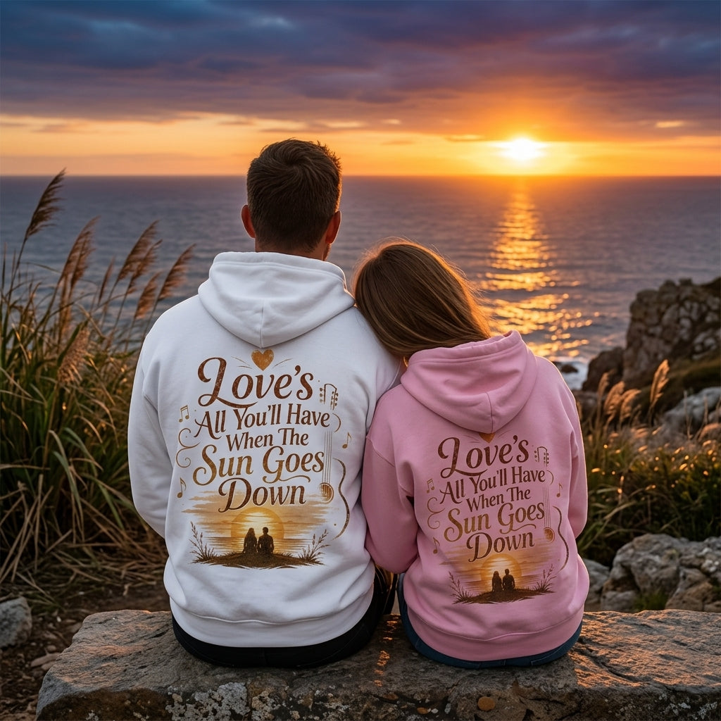Couple wearing matching white and pink hoodies printed with sunset lyric graphic, sitting by the ocean at sunset