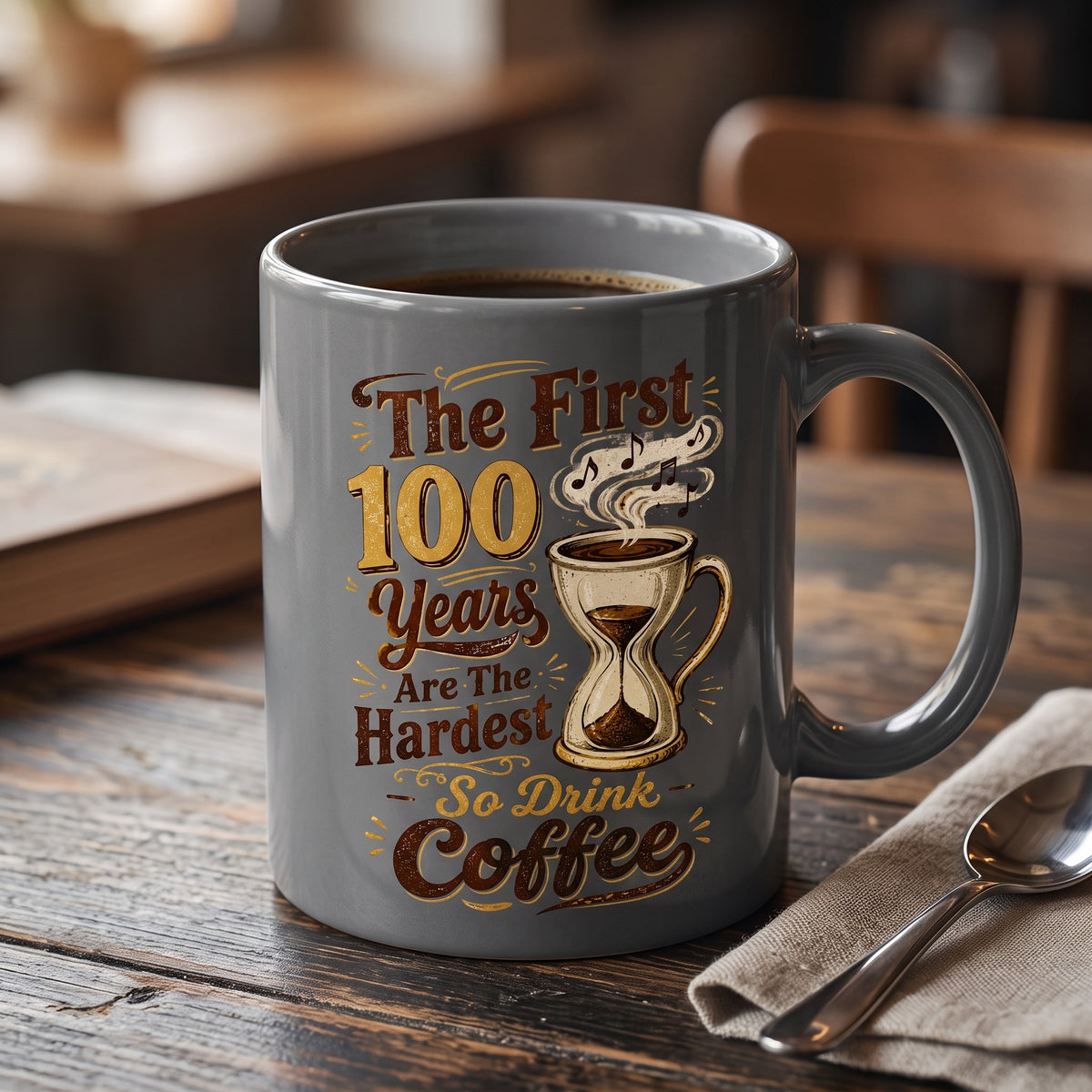 Gray ceramic mug with brown and gold text reading "The First 100 Years Are The Hardest So Drink Coffee"